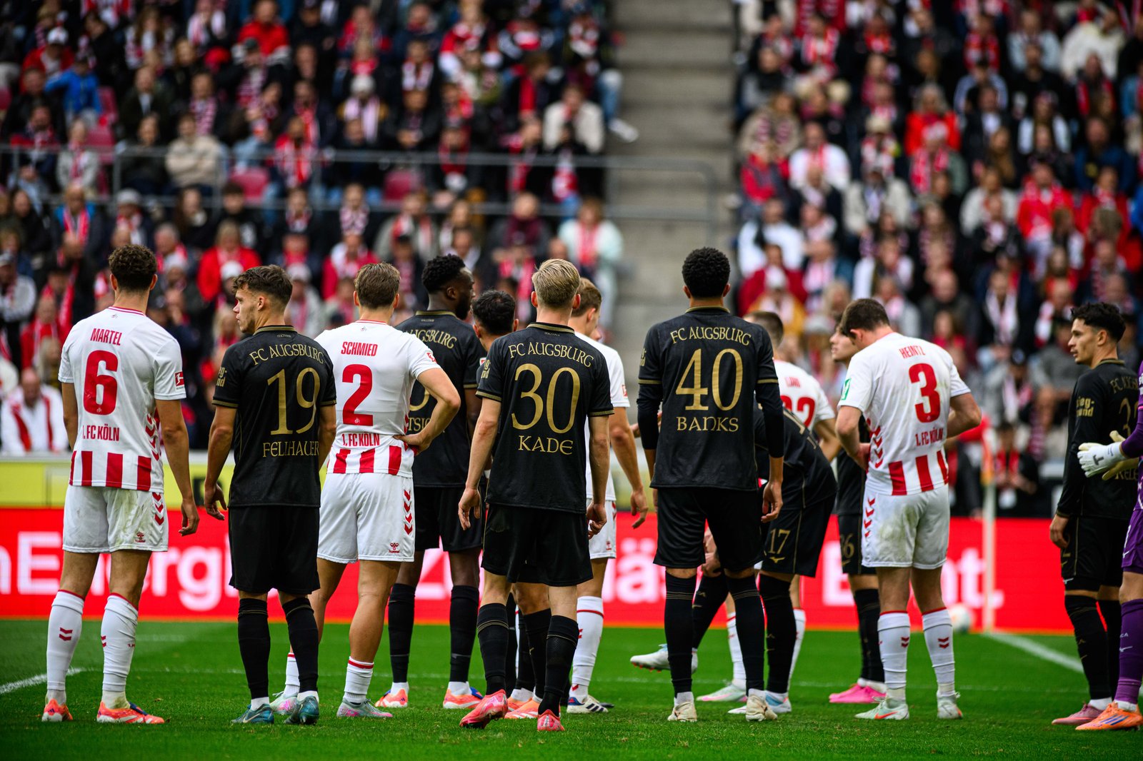 Spieler von 1. FC Köln und FC Augsburg stehen sich beim Bundesliga-Duell im Rhein-Energie-Stadion in Köln gegenüber. Beim Aufeinandertreffen am 18. Oktober 2025 sind die Augsburger in ihren schwarzen Auswärtstrikots mit den Nummern 19 Fellhauer, 30 Kade und 40 Banks gut zu erkennen, während die Kölner in ihren rot-weißen Heimtrikots daneben aufgereiht stehen. (Copyright Depositphotos.com)