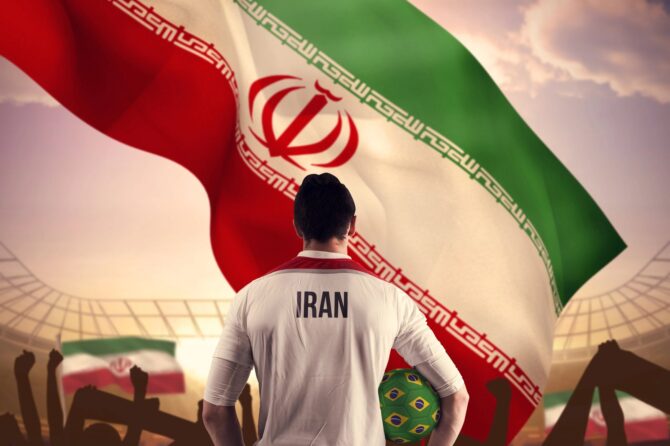 Ein Fußballspieler im Trikot der iranischen Nationalmannschaft steht mit einem Fußball in der Hand vor einer großen iranischen Fahne, im Hintergrund jubelnde Fans im Stadion. (Copyright Depositphotos.com)
