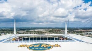 Blick vom Dach des Hard Rock Stadium in Miami Gardens, Florida – Austragungsort der FIFA Fußball-Weltmeisterschaft 2026. Das Hard-Rock-Logo ist auf dem weitläufigen weißen Zeltdach gut zu erkennen, dahinter erstreckt sich die flache Stadtlandschaft Miamis unter einem dramatischen Wolkenhimmel. (Copyright Depositphotos.com)