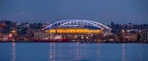 Das Lumen Field in Seattle erstrahlt in der Abenddämmerung – aufgenommen am 17. Oktober 2025 vom Puget Sound aus. Das erleuchtete Stadion mit seinem markanten Bogendach spiegelt sich im Wasser, im Vordergrund liegt ein Küstenwachschiff am Kai. Das Lumen Field ist einer der Austragungsorte der FIFA Fußball-Weltmeisterschaft 2026. (Copyright Depositphotos.com)