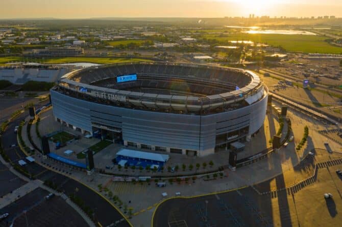 Luftaufnahme des MetLife Stadium in East Rutherford, New Jersey, bei Sonnenuntergang. Die Drohnenaufnahme zeigt das imposante Stadion mit dem umlaufenden MetLife-Schriftzug von außen, eingebettet in die flache Landschaft New Jerseys mit der Skyline von Manhattan am Horizont. Das MetLife Stadium ist Austragungsort der FIFA Fußball-Weltmeisterschaft 2026. (Copyright Depositphotos.com)
