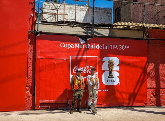 Zwei Bauarbeiter stehen vor einem roten Werbeplakat des WM-2026-Sponsors Coca-Cola nahe des Aztekenstadions in Mexiko-Stadt - eine Szene aus der ZDF-Reportage "Greenwashed? - FIFA WM 2026: Gruen ist hier nur der Rasen". ZDF/Svaantje Schroeder