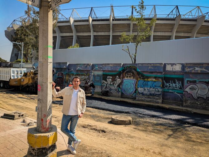ZDF-Reporter Sherif Rizkallah steht auf einer unbefestigten Strasse direkt vor dem Aztekenstadion in Mexiko-Stadt, im Hintergrund Baufahrzeuge - aus der Reportage "Greenwashed? - FIFA WM 2026: Gruen ist hier nur der Rasen". ZDF/Svaantje Schroeder
