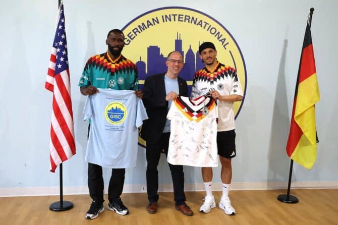 Leroy Sané (rechts) und Antonio Rüdiger (links) vom deutschen Nationalteam posieren bei einem Meet-and-Greet mit Schulleiter Rasmus Frederich an der German International School Chicago am 4. Juni 2026 in Chicago. Sané präsentiert dabei das legendäre weiße DFB-Trikot von 1994, während Rüdiger im bunten Auswärtstrikot der Neunzigerjahre auftritt. Die beiden Profis besuchten die zweisprachige deutsch-amerikanische Schule während der WM-Vorbereitung. (Copyright AFP/Alexander Hassenstein)