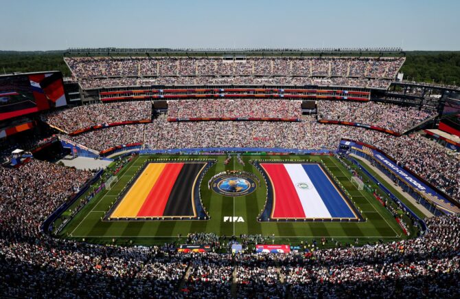 Riesige Nationalflaggen von Deutschland und Paraguay bedecken den Rasen, während sich beide Mannschaften und das Schiedsrichtergespann vor dem Anpfiff im prall gefüllten Boston Stadium aufstellen. Aufgenommen vor dem Sechzehntelfinale der WM 2026 zwischen Deutschland und Paraguay in Foxborough am 29. Juni 2026. (Darrian Traynor / Getty Images)