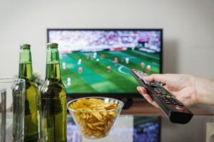 Eine Hand mit Fernbedienung vor einem Fernseher mit Fußballspiel, davor zwei Bierflaschen und eine Schale Chips