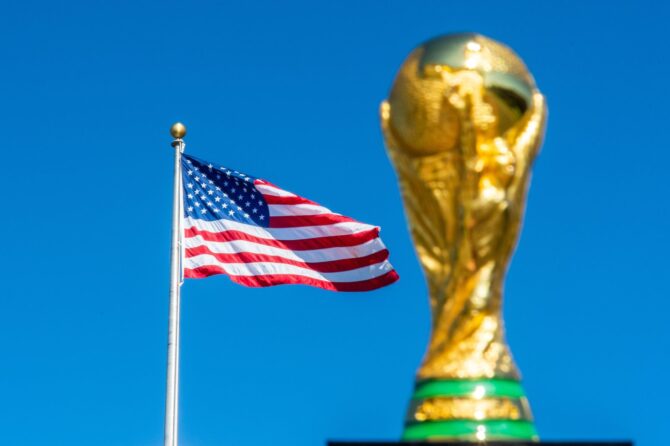 Die US-amerikanische Flagge weht vor blauem Himmel, daneben ragt der WM-Pokal empor. USA ist einer von drei Ausrichter der FIFA Fußball-Weltmeisterschaft 2026. (Copyright Depositphotos.com)