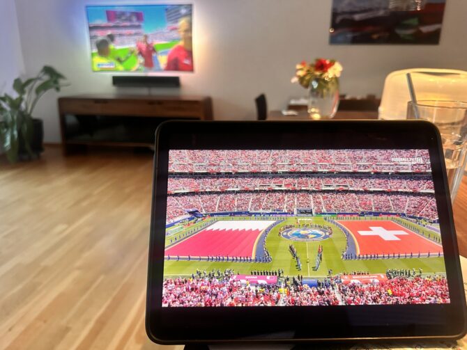 Auf einem Tablet im heimischen Wohnzimmer läuft die Übertragung des WM-2026-Gruppenspiels der Gruppe B zwischen der Schweiz und Katar: Vor dem Anpfiff werden die übergroßen Flaggen beider Nationen auf dem Rasen ausgebreitet, während die Mannschaften und Schiedsrichter im Mittelkreis aufgereiht stehen. Im Hintergrund zeigt auch der Fernseher das Spiel.