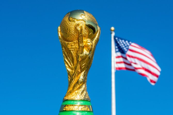 Eine Nachbildung des WM-Pokals vor strahlend blauem Himmel, im Hintergrund weht die Flagge der USA. Die Vereinigten Staaten richten 2026 gemeinsam mit Mexiko und Kanada die FIFA Fußball-Weltmeisterschaft aus. (Copyright Depositphotos.com)