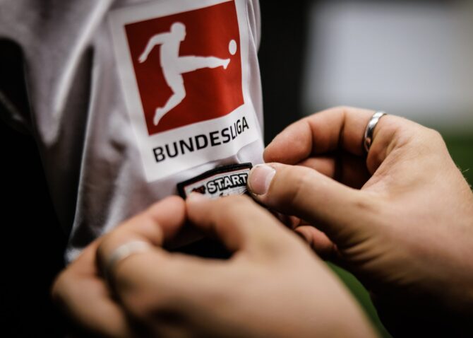Der Zeugwart drückt das Startelfdebüt-Patch auf den Ärmel eines Bundesliga-Trikots