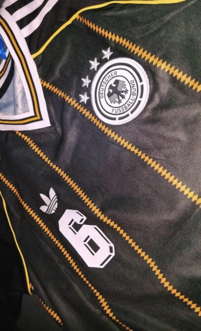 Detailaufnahme des adidas-DFB-Abschiedstrikots 2026: historisches DFB-Wappen mit vier Sternen, adidas-Trefoil und goldene Zickzack-Nadelstreifen auf schwarzem Grund. (Bild: Footy Headlines)